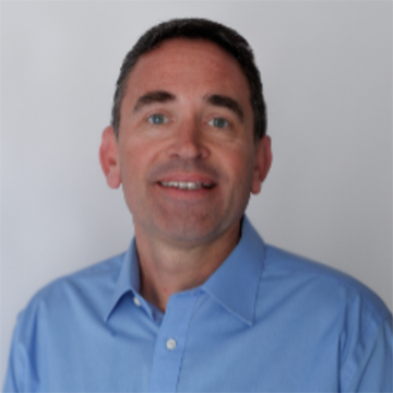 A man with short dark hair and blue eyes smiles at the camera. He is wearing a light blue shirt and has a neutral background.