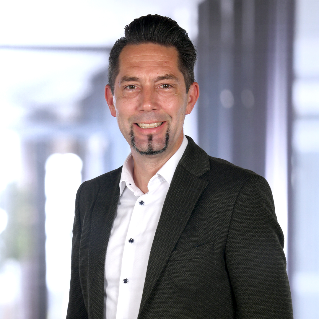 A man smiling confidently while wearing a dark blazer over a white shirt. The background features blurred office-like elements, suggesting a professional setting.