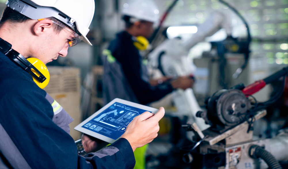 A factory worker in a hard hat and headphones monitors machinery using a tablet, while another worker operates a robotic arm in the background.
