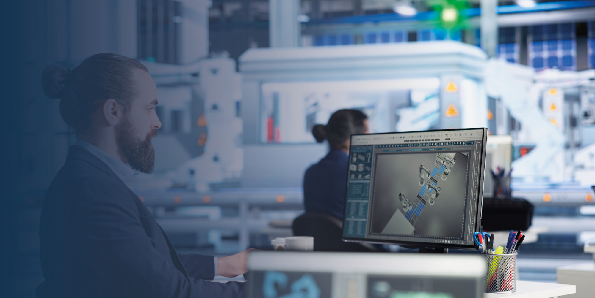 A man with a beard sits at a desk, focused on a computer screen displaying a 3D design in a modern workspace. In the background, another person works at a separate station amid an industrial setting with machinery.