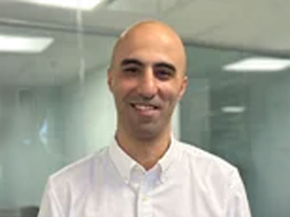 A smiling bald man wearing a white button-up shirt stands in an office setting with glass walls in the background.