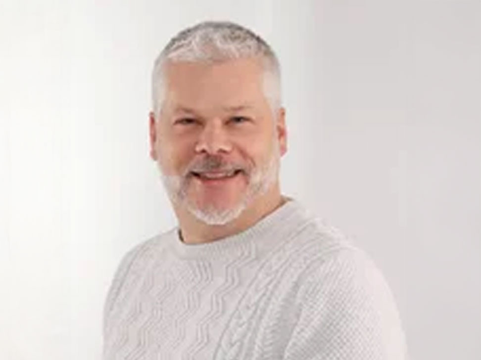 Ein lächelnder Mann mit grauem Bart und kurzen grauen Haaren trägt einen weißen, strukturierten Pullover. Der Hintergrund ist hell und neutral.