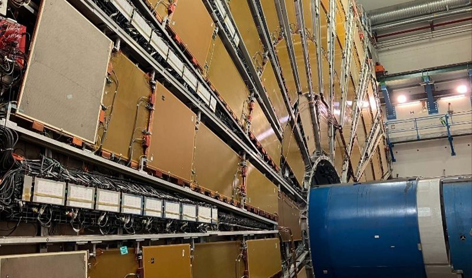 A large cylindrical structure with intricate wiring and electronic panels, set against a wall in a research facility. The structure appears to be part of a scientific experiment, likely a particle accelerator or detector.