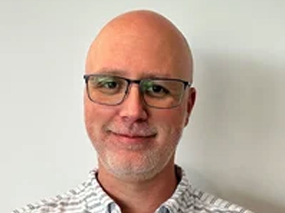 A man with a shaved head and glasses smiles at the camera, wearing a collared shirt with a checkered pattern.