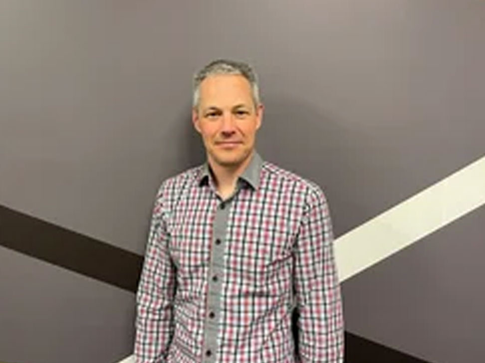 A man stands smiling in front of a gray wall with a white and brown geometric design. He is wearing a checked shirt with a button-up style and has short, light-colored hair.