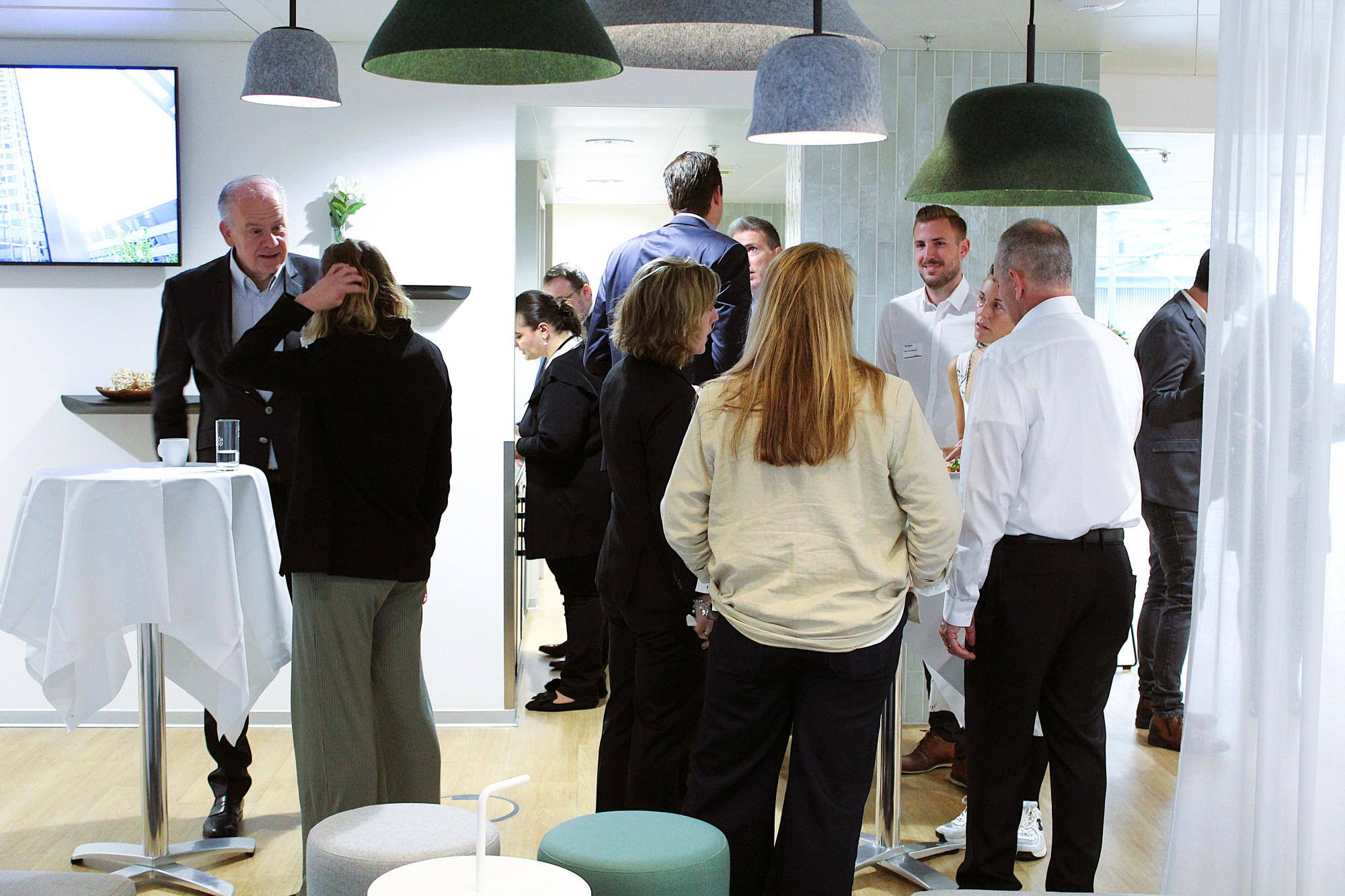 A group of professionals engaged in conversation at a networking event in a modern, brightly lit space. Several people are standing around tables, while others are gathered in small groups. The setting features stylish lighting and comfortable seating.