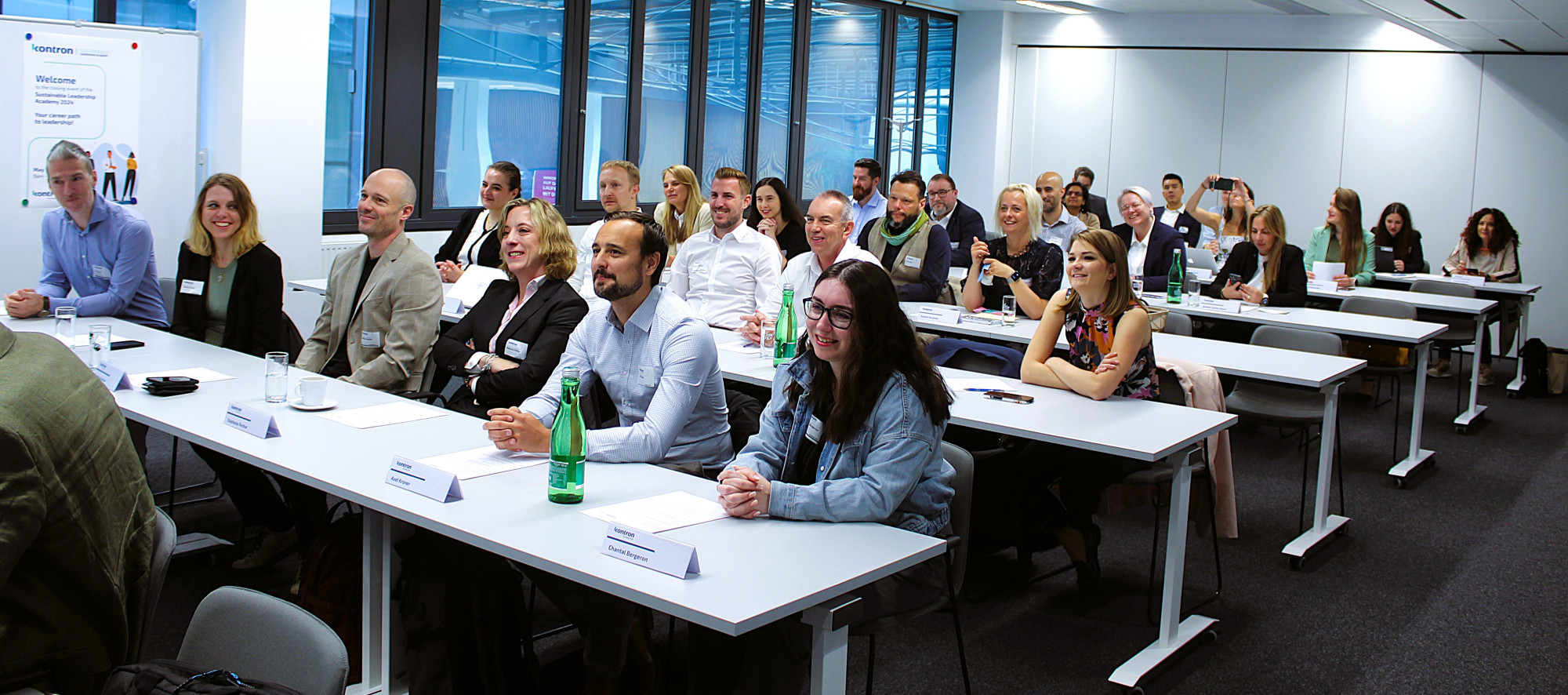 A diverse group of people seated in a modern conference room, attentively listening to a presentation. They are arranged in rows at long tables, with some taking notes while others engage with one another. Bright overhead lighting and large windows illuminate the space.