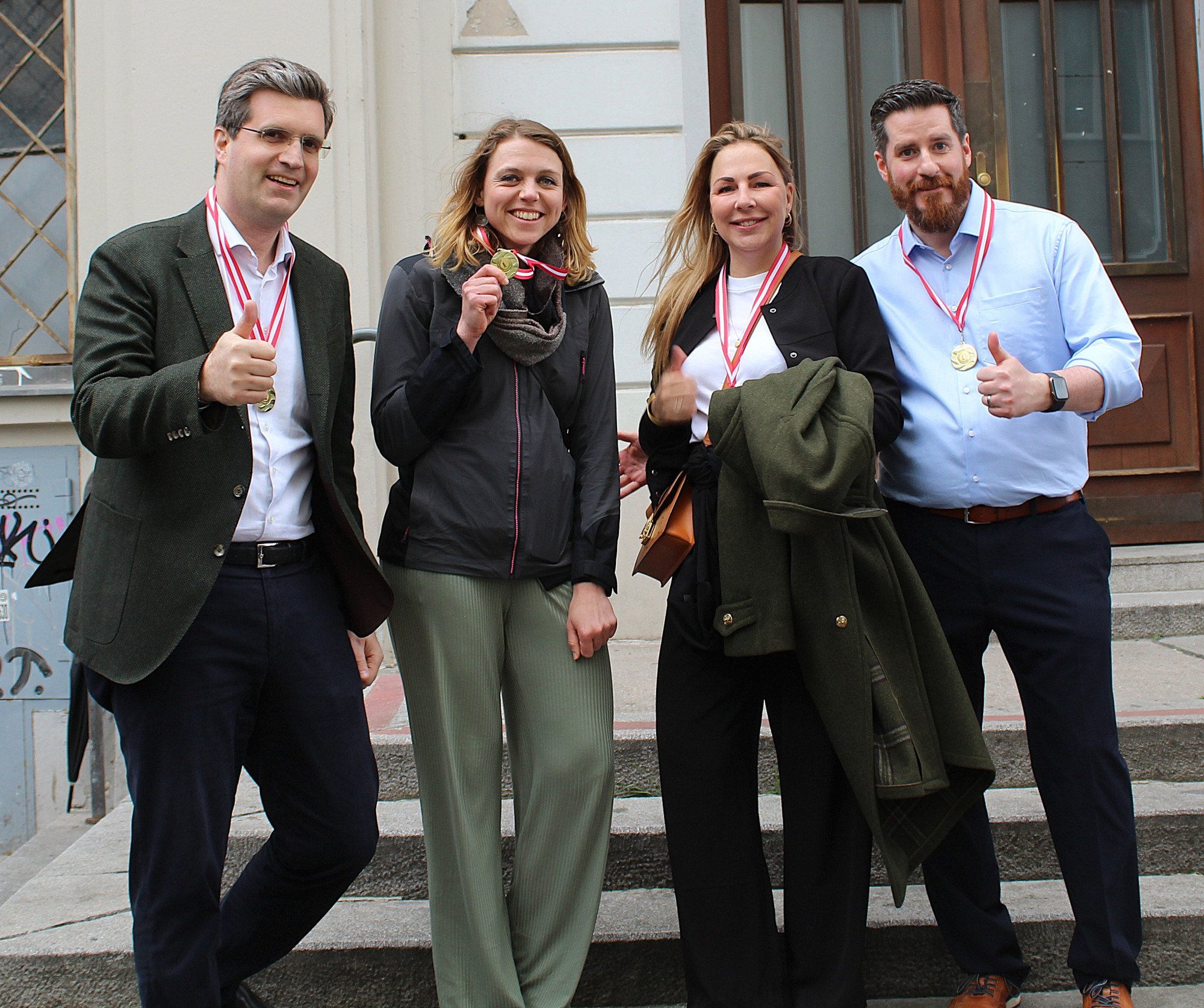 Four smiling individuals pose together on a set of steps, all wearing medals around their necks. They give thumbs-up gestures, dressed in casual yet stylish clothing, with a mix of winter and spring attire. The background includes a building facade.