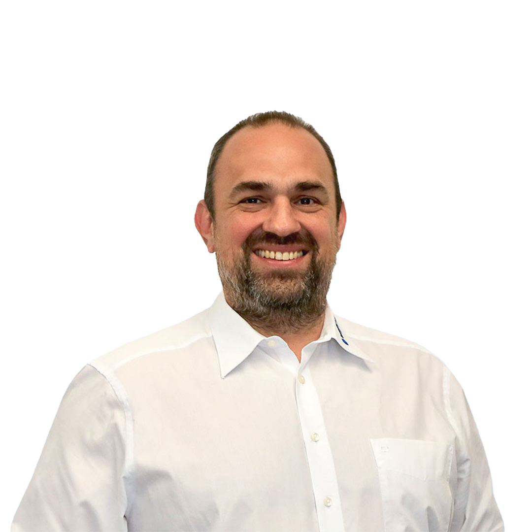 A smiling man with a beard stands against a white background, wearing a light blue collared shirt.