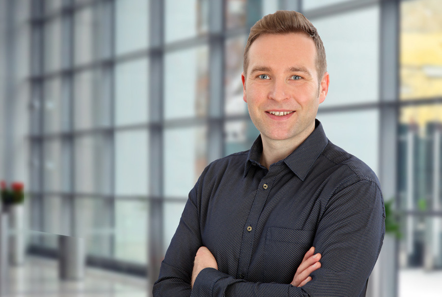 A smiling man with short brown hair stands with his arms crossed in a modern, glass-walled environment. He wears a dark, button-up shirt and is positioned against a blurred background that suggests a bright, airy space.