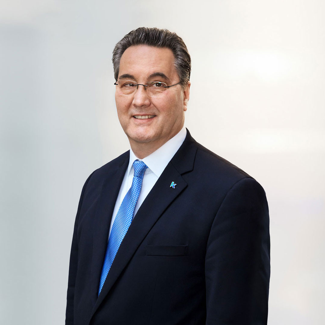 Ein Mann in einem dunklen Anzug mit blauer Krawatte und Brille lächelt freundlich in die Kamera. Der Hintergrund ist hell und unscharf.