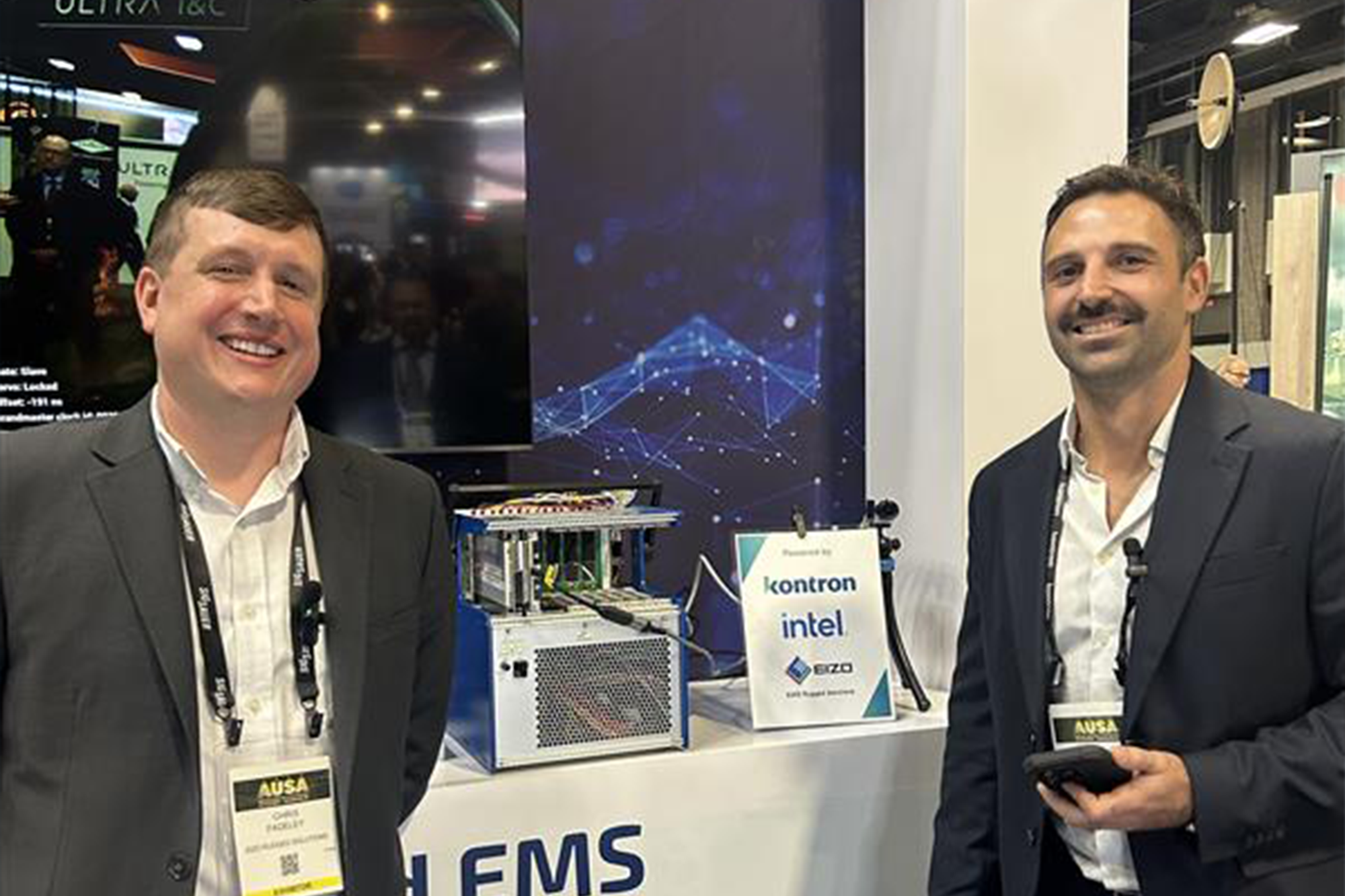 Two men in suits stand smiling in front of a display showcasing a technology product. The background features a screen with digital graphics and banners, with a logo for Kontron and Intel visible.