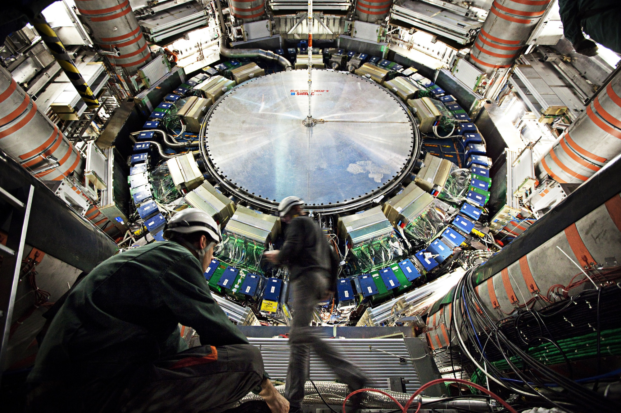 An expansive view inside a large scientific facility, featuring a circular structure with various machines and cables. Two workers in helmets are seen, one sitting on a step and the other walking across the floor. The environment is industrial and high-tech, indicating a complex research or experimental setup.