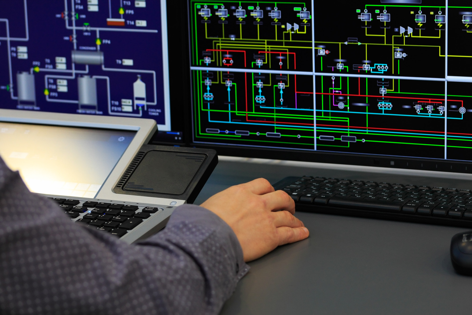 A person’s hand operates a laptop and is positioned near a computer keyboard, while multiple digital screens display intricate electrical diagrams with colorful lines representing connections and signals.