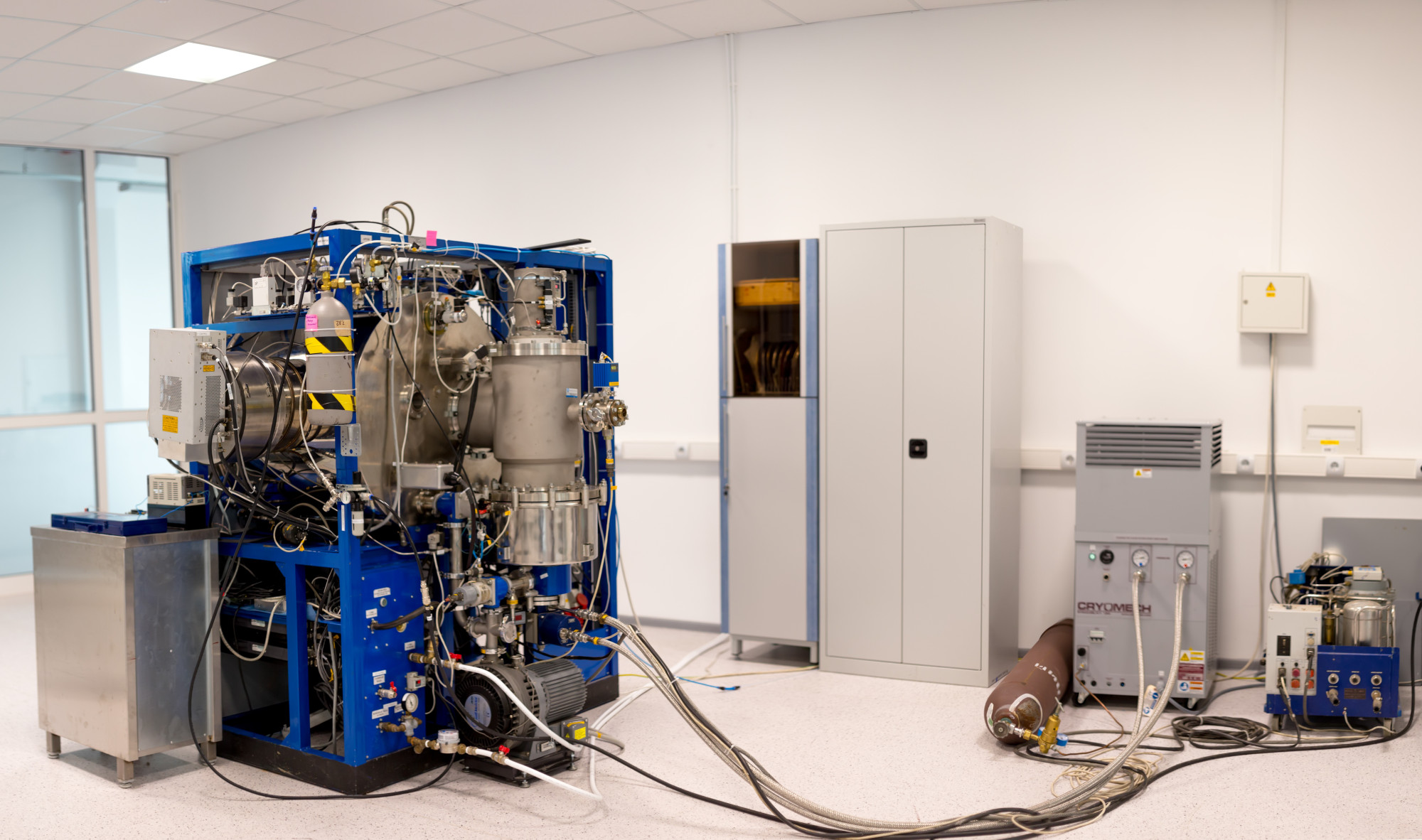 A large, complex machine with various tubes and components occupies one side of a clean, well-lit laboratory. Nearby, a gray storage cabinet and a smaller device with a digital screen are visible. The floor is light-colored, and the room has plain white walls.