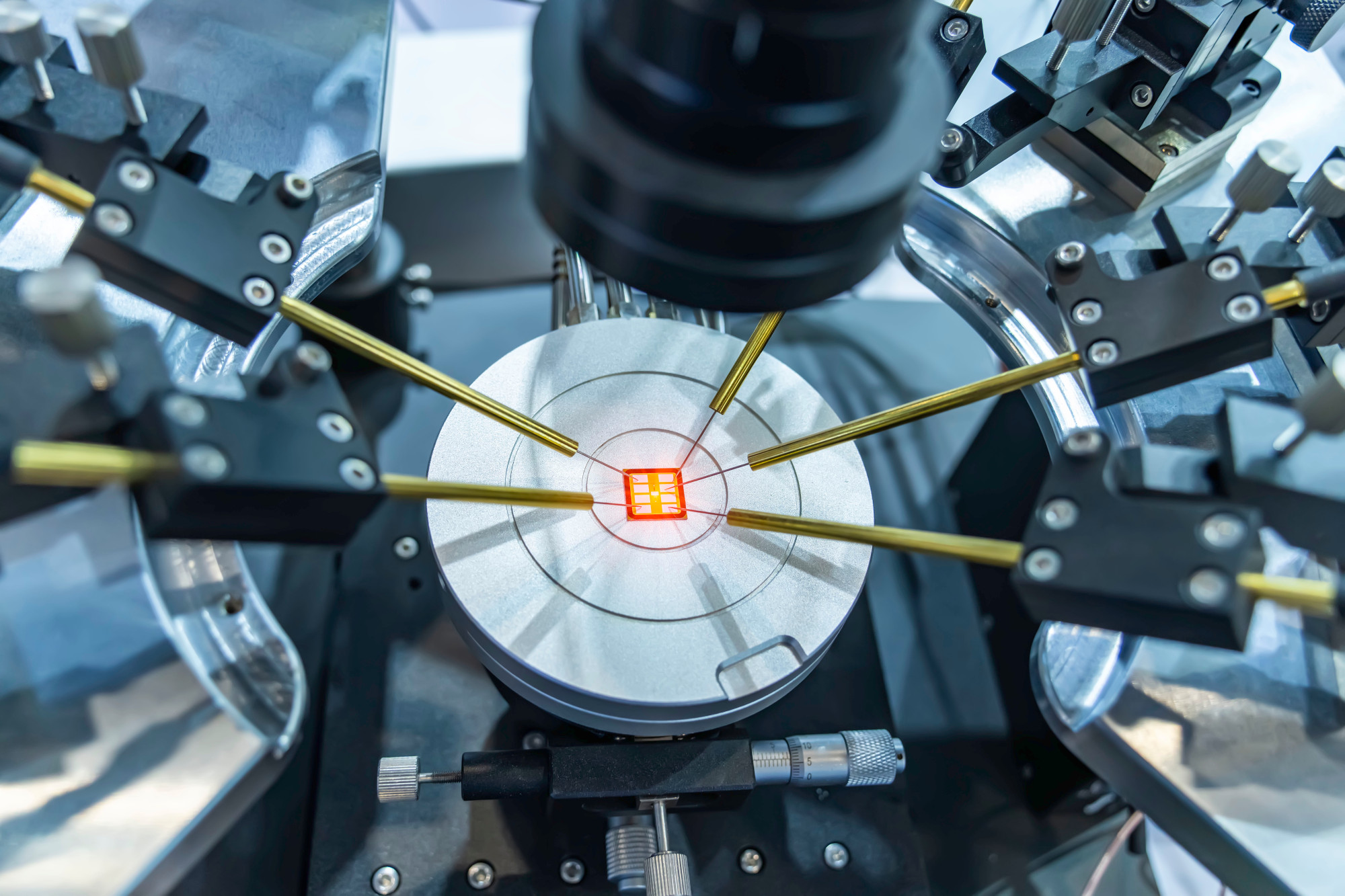 A close-up view of a high-tech device used for semiconductor testing, featuring multiple gold probes positioned over a circular base with a lit red square in the center.