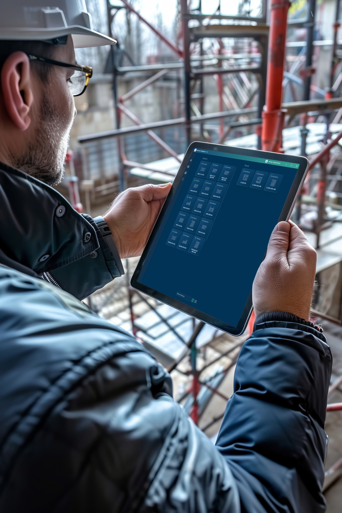A construction worker wearing a hard hat and glasses holds a tablet, looking at a digital interface while standing in a building site surrounded by scaffolding.