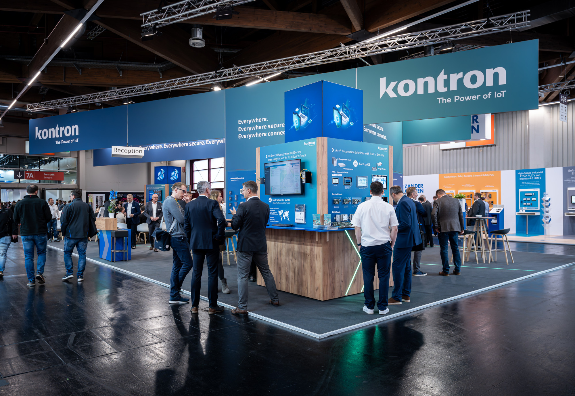 A trade show booth for Kontron, featuring large blue signage and various interactive displays. Attendees engage at the booth with several standing tables and a reception area in the background.