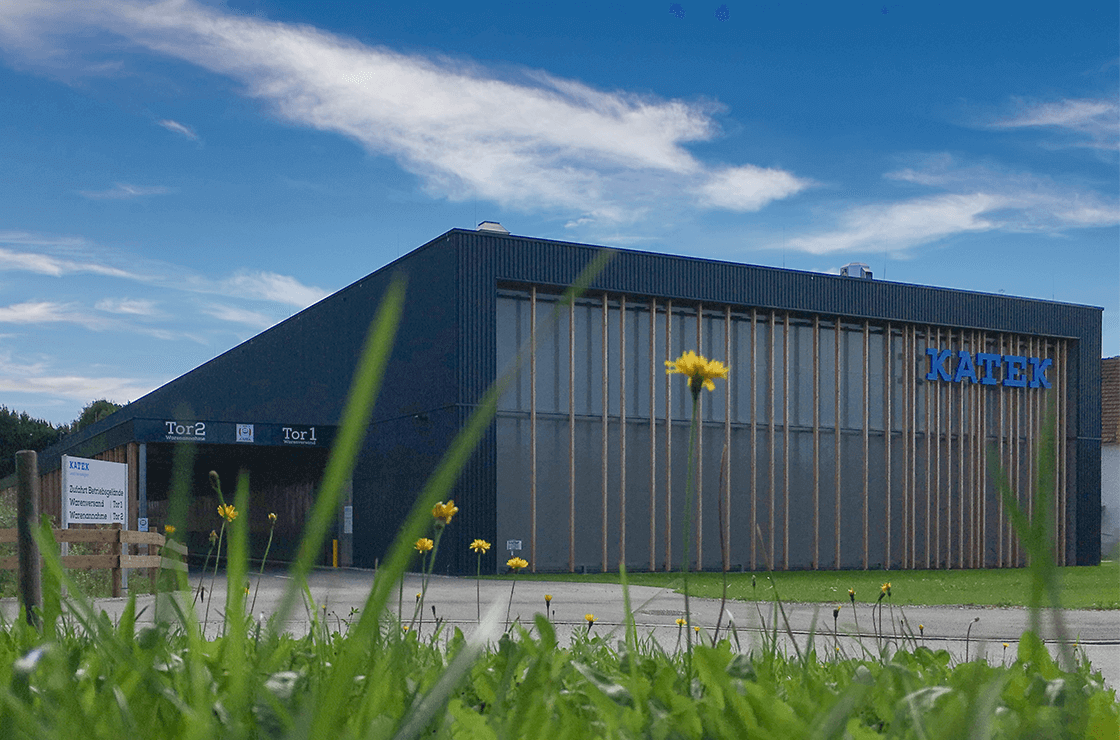 A modern industrial building with a blue "KAUTEK" sign. The structure features large windows and wooden accents, set against a clear sky. In the foreground, grass and yellow dandelions are visible.