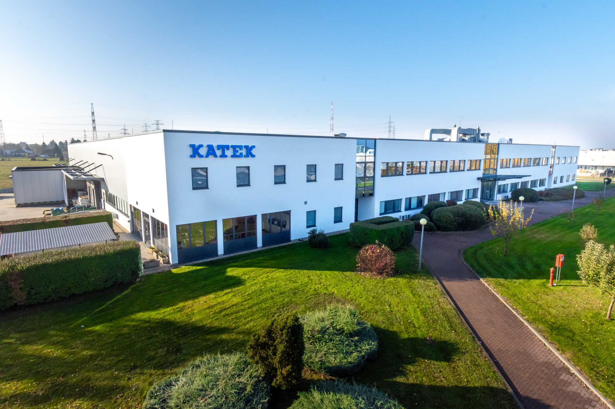 A modern white industrial building with the word "Katek" prominently displayed on the exterior. Surrounding greenery includes neatly trimmed hedges and a well-maintained lawn. The scene is clear against a blue sky.