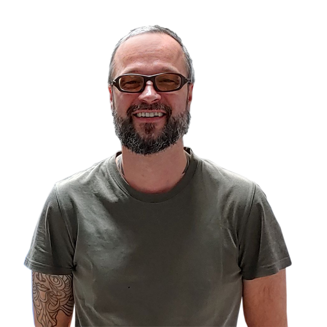A smiling man with a beard and glasses wearing a gray t-shirt stands against a white background. He has visible tattoos on his right arm.