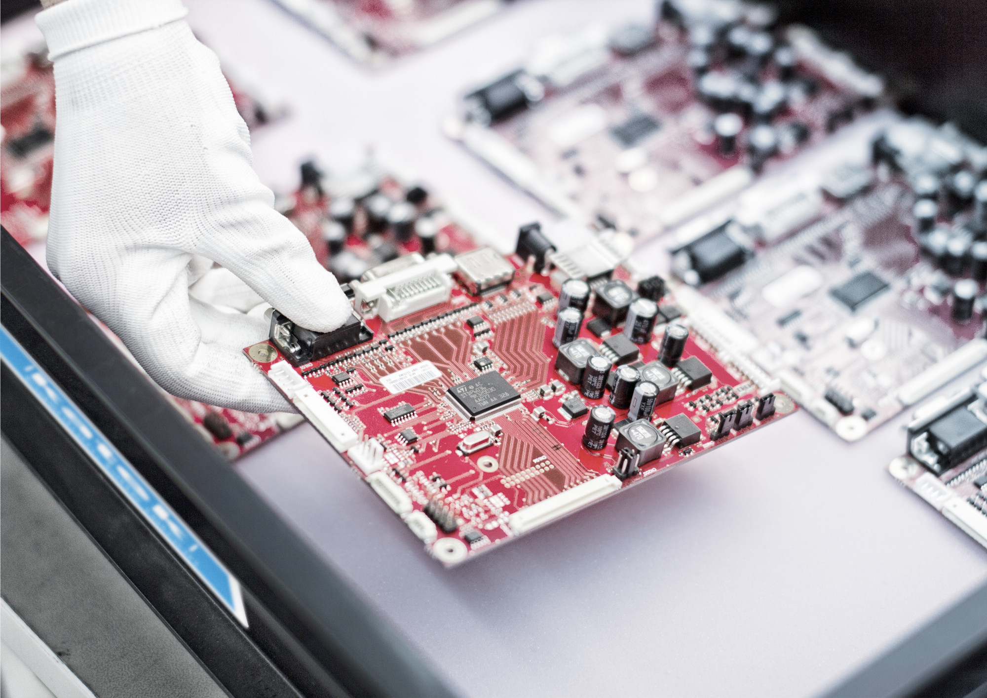 A gloved hand holds a red circuit board with various components, surrounded by other circuit boards on a workspace.