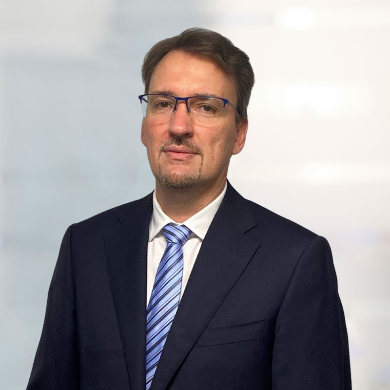A man in a dark suit and blue tie stands against a soft, blurred background. He has short brown hair and wears rectangular glasses, looking confidently at the camera.