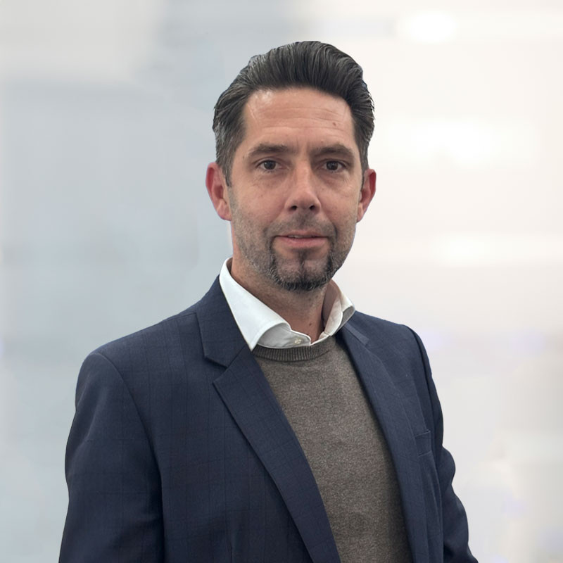 A man with short, dark hair and a trimmed beard is wearing a navy blazer over a light brown sweater. He stands confidently, with a neutral background suggesting a professional setting.