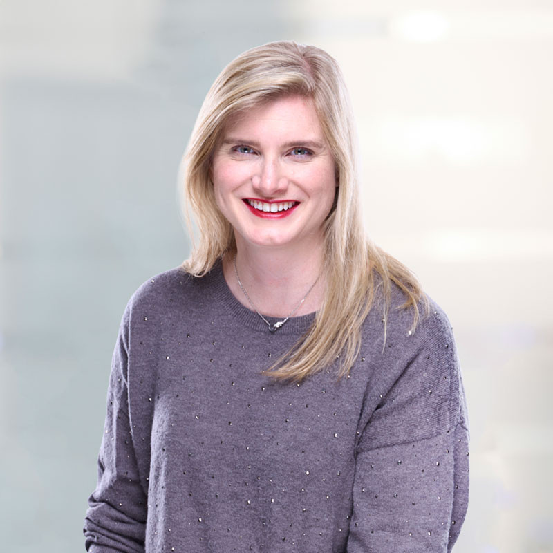 A smiling woman with long blonde hair and red lipstick is wearing a gray sweater. She is posed against a light, blurred background.