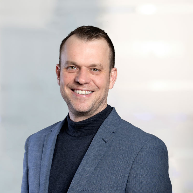 A man with short hair and a friendly smile wears a gray blazer over a black turtleneck. He stands against a light background, projecting a professional demeanor.