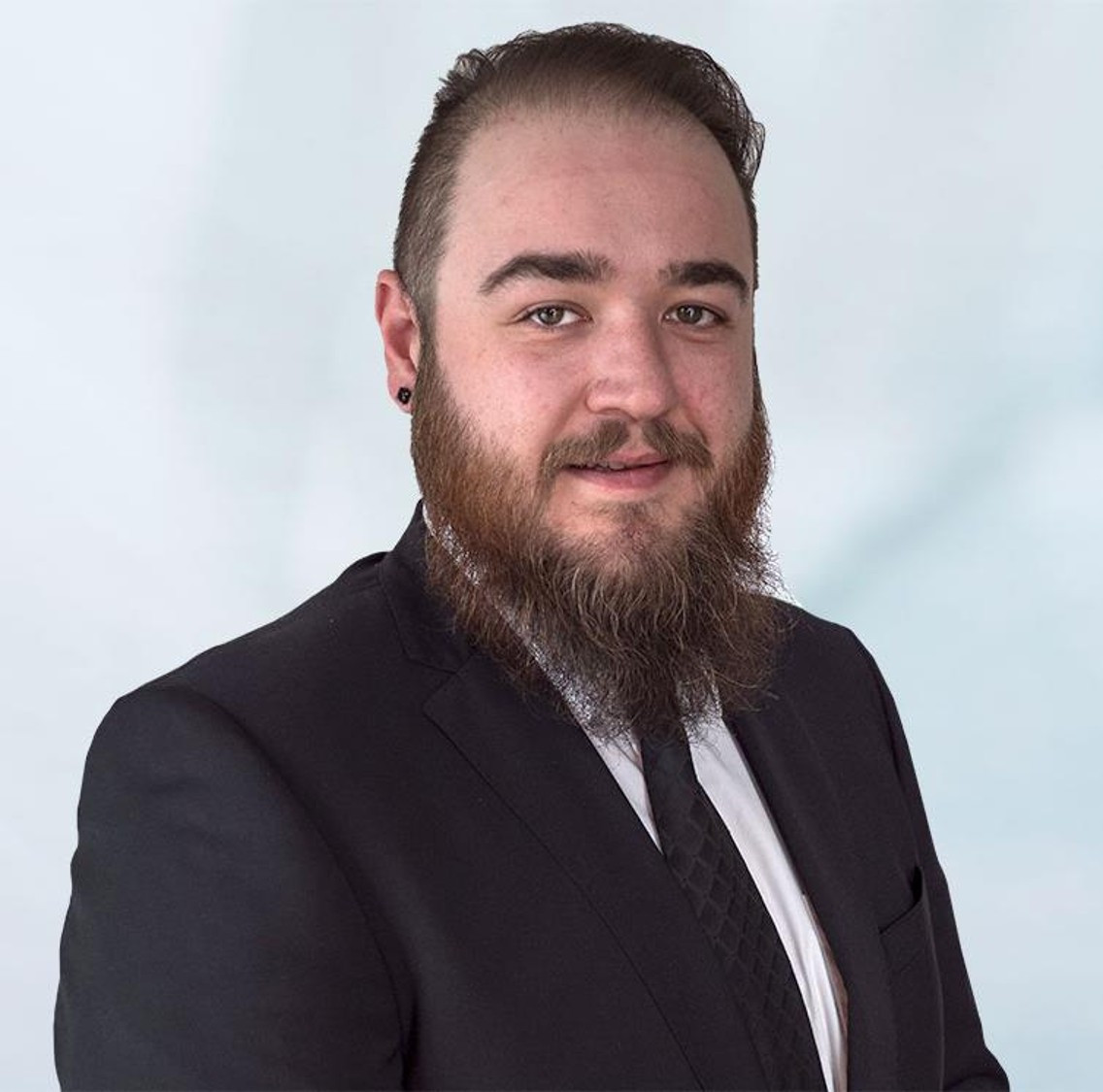 A man with a thick beard and short hair is wearing a black suit with a white shirt and black tie. He has a slight smile and is positioned against a light blue background.