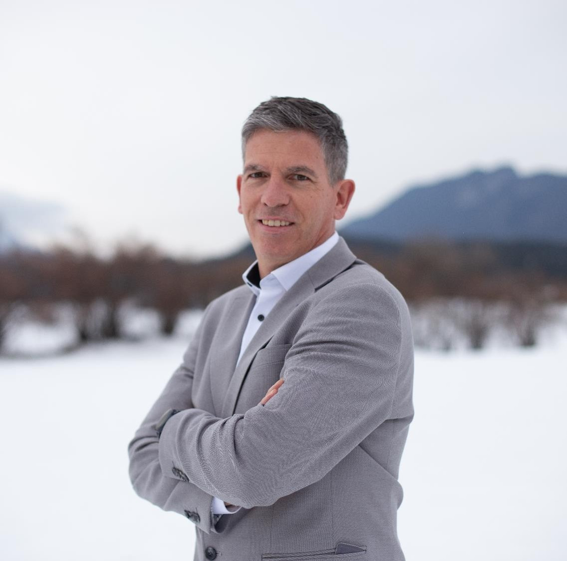 A man smiles confidently while standing outdoors in a snowy landscape. He is wearing a light gray suit jacket and has his arms crossed. Mountains are visible in the background under a cloudy sky.