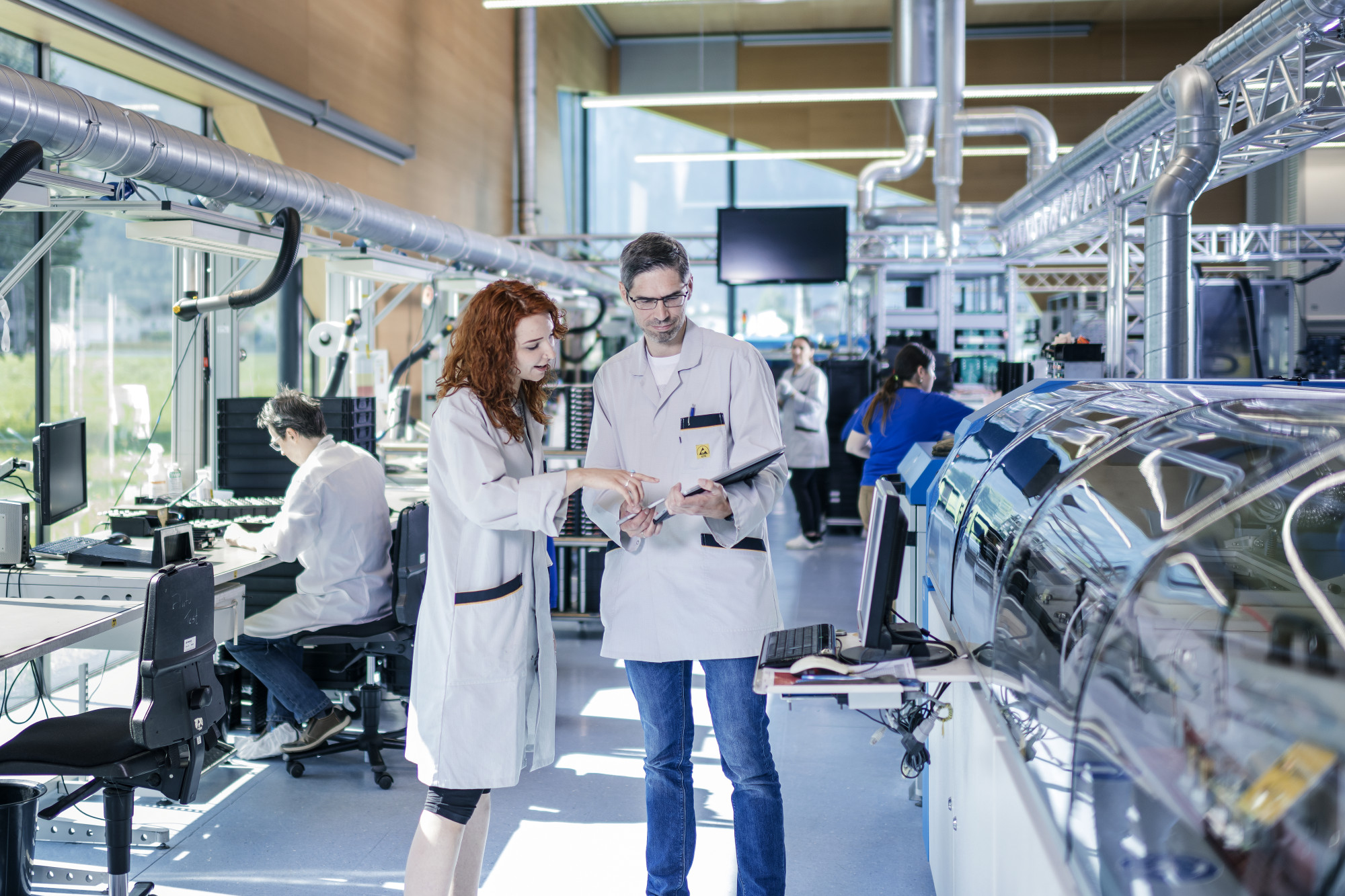 Two scientists in lab coats discuss findings in a modern laboratory. One woman points to a tablet while the man observes. In the background, additional researchers work at various stations. Bright, natural light floods the space from large windows.