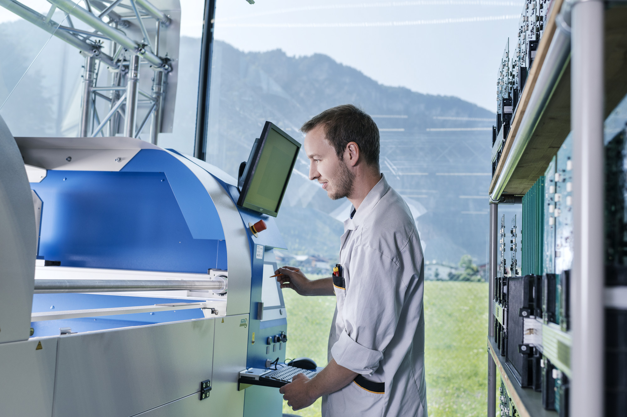 A technician operates a blue and white machine in a bright workshop, with a mountain landscape visible through large windows in the background. The technician wears a lab coat and focuses on the machine's control panel.