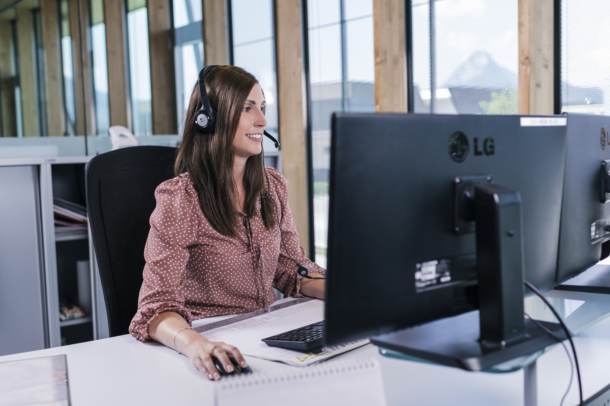 A woman with shoulder-length brown hair is sitting at a desk wearing a headset. She is smiling while looking at a computer monitor, with paperwork and a notepad in front of her. The workspace features large windows with natural light.