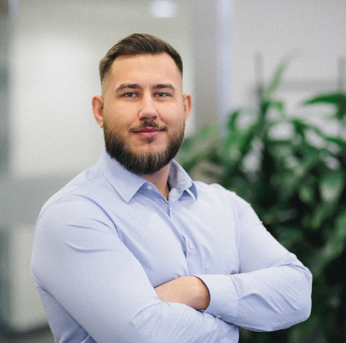 A man with a beard and short hair is standing with his arms crossed, wearing a light blue shirt. He is smiling confidently in an indoor setting with green plants in the background.