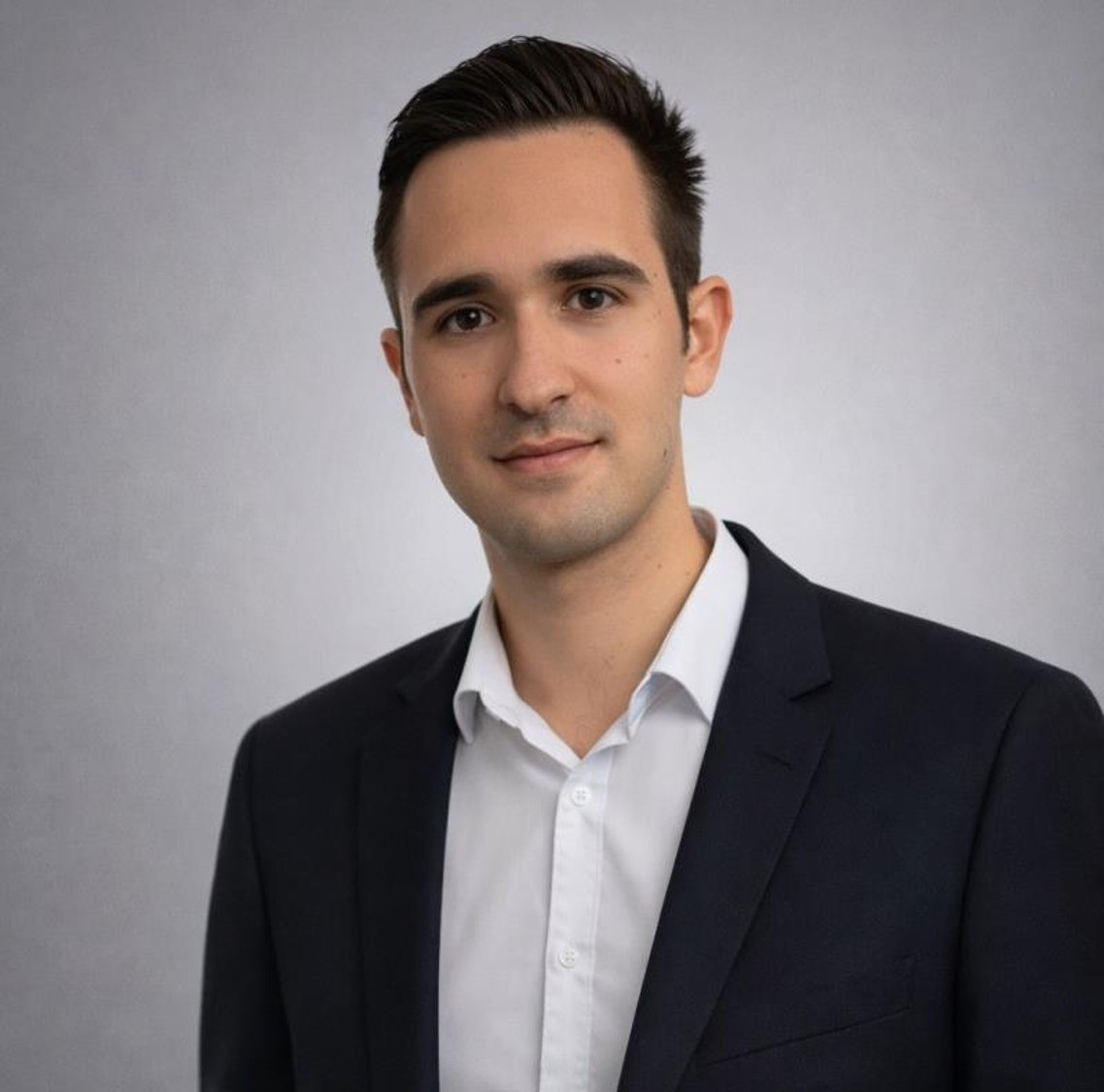 A young man in a dark suit and white shirt stands against a neutral background, looking directly at the camera with a slight smile.