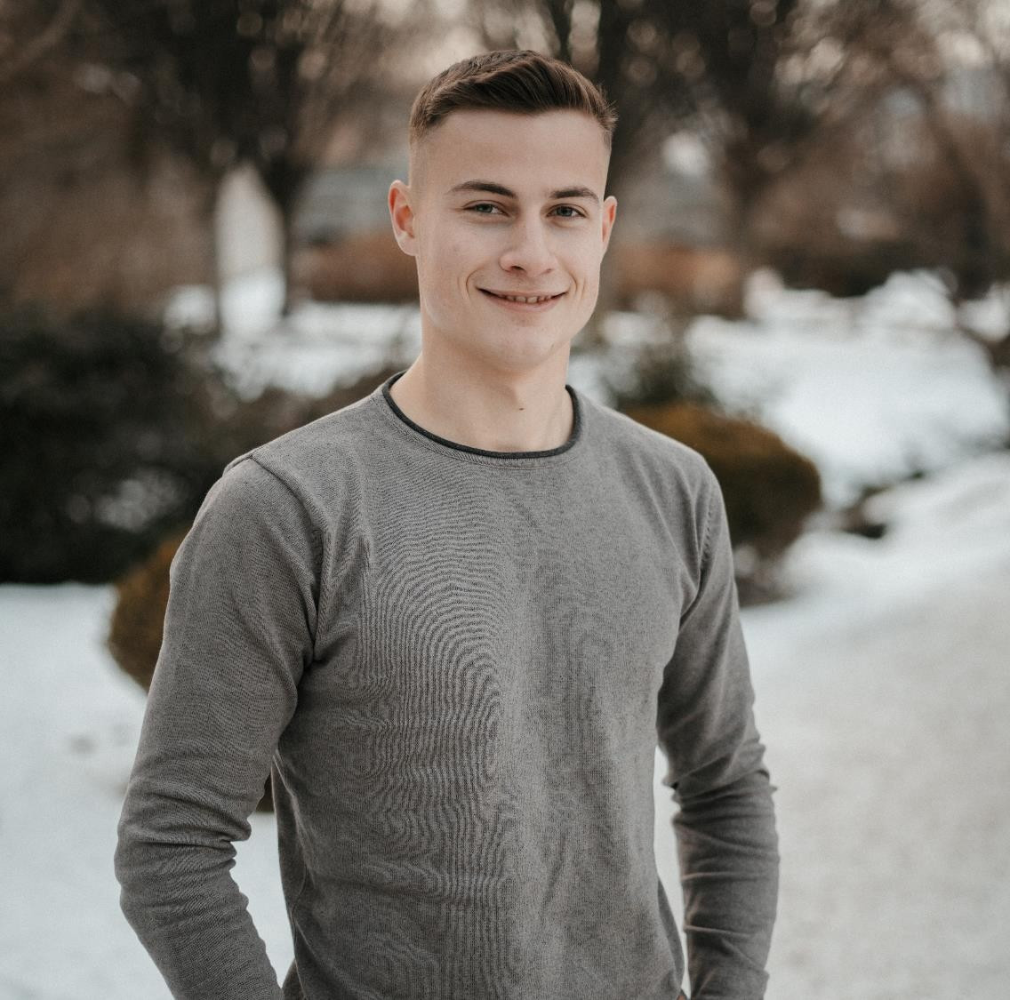 A young man stands outdoors, smiling confidently. He is wearing a long-sleeved gray shirt, with snow and trees visible in the background. The scene suggests a winter setting.