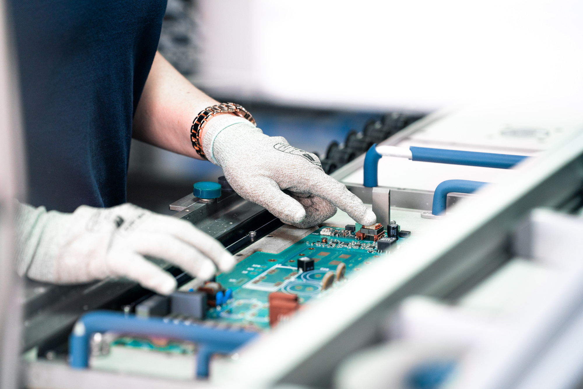 A person wearing protective gloves is working on a circuit board, examining components and connections. The background shows machinery and tools related to electronic assembly.