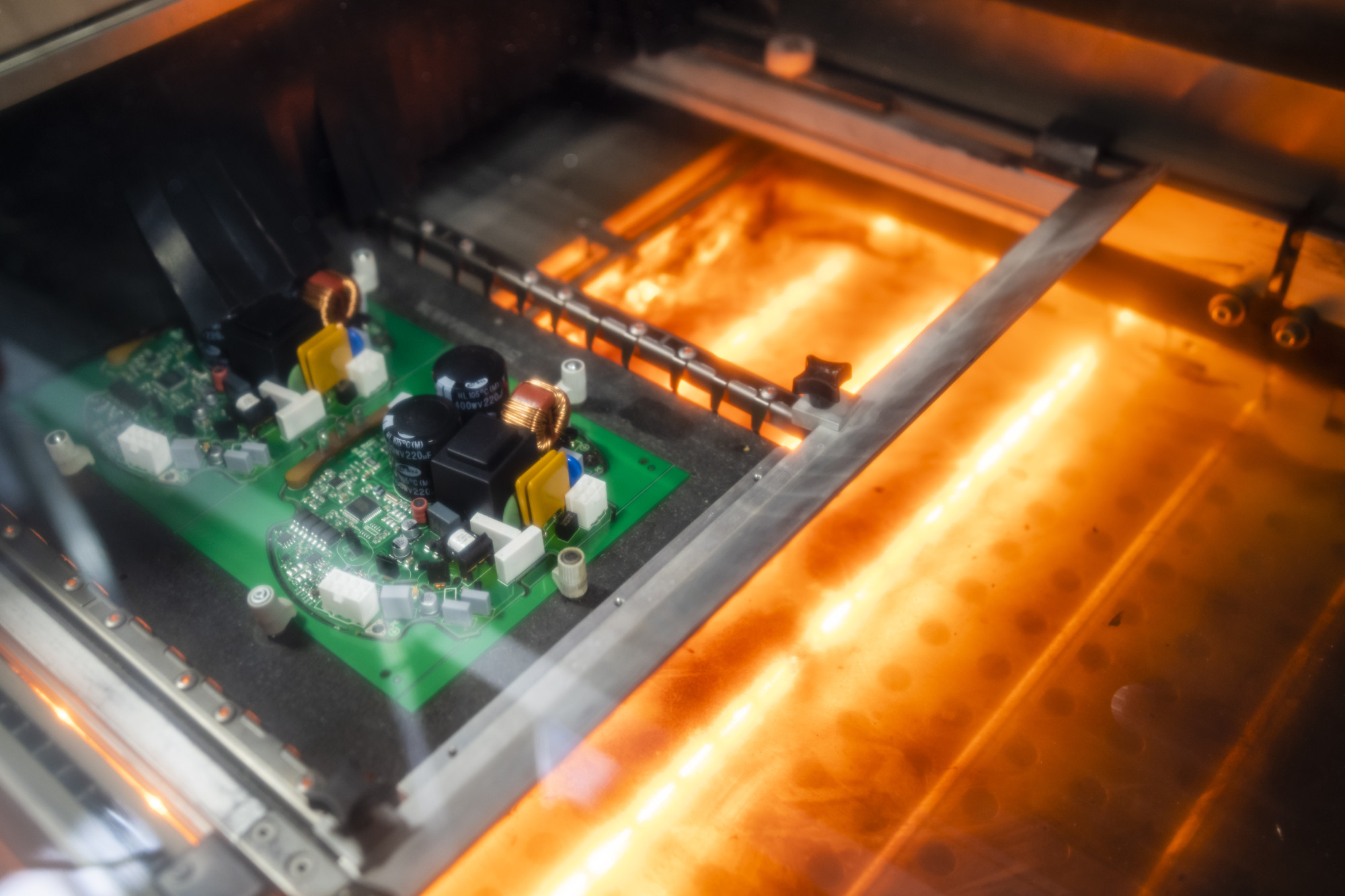 A close-up view of two green circuit boards inside a machine, illuminated by warm orange light. The boards contain various electronic components like capacitors and inductors.