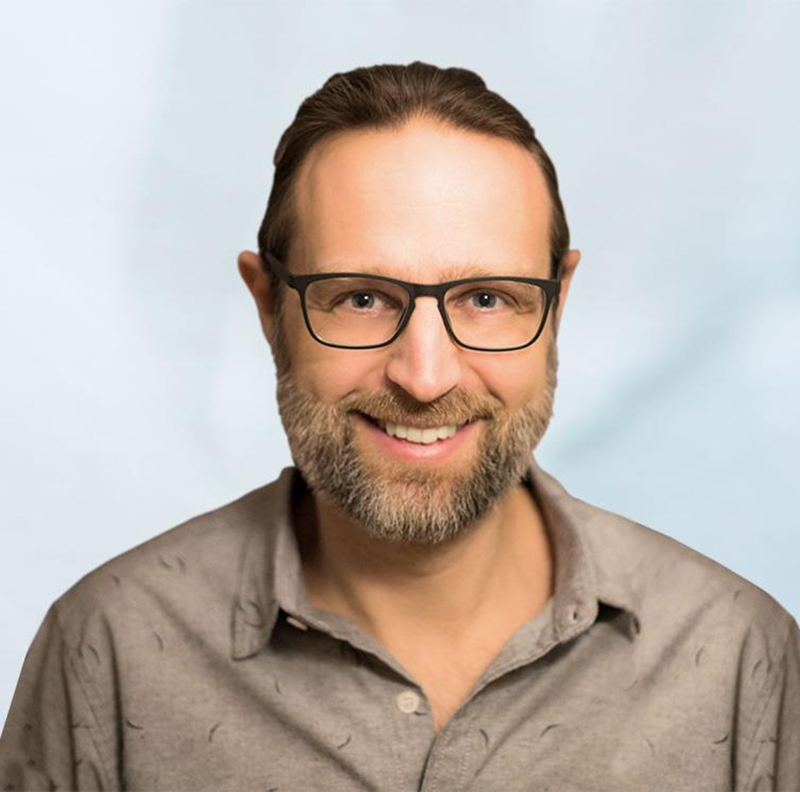 A smiling man with glasses and a beard wearing a light gray shirt is facing the camera against a soft, light blue background.
