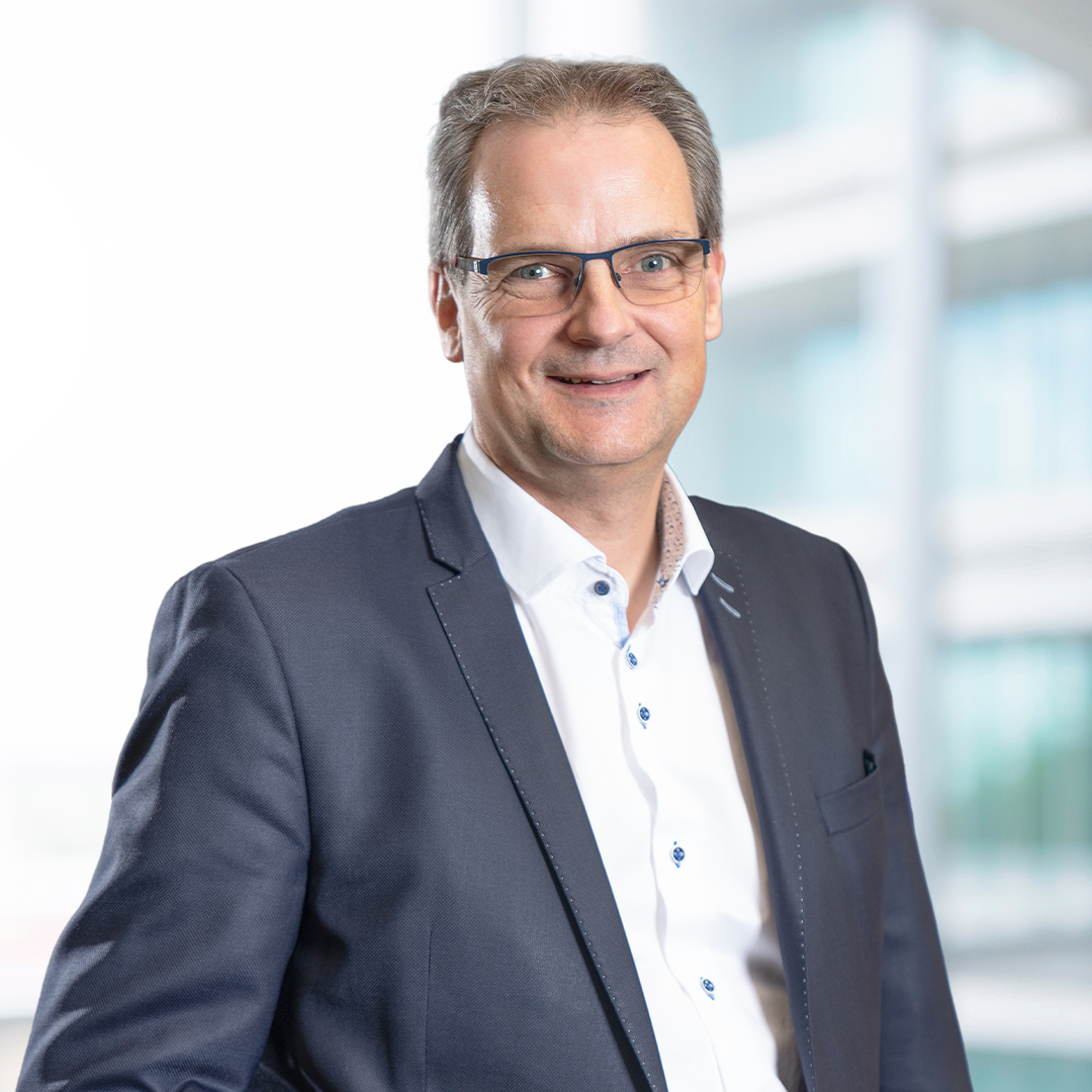 A man wearing glasses smiles confidently while wearing a dark blazer over a white shirt. The background features a softly blurred modern office setting.