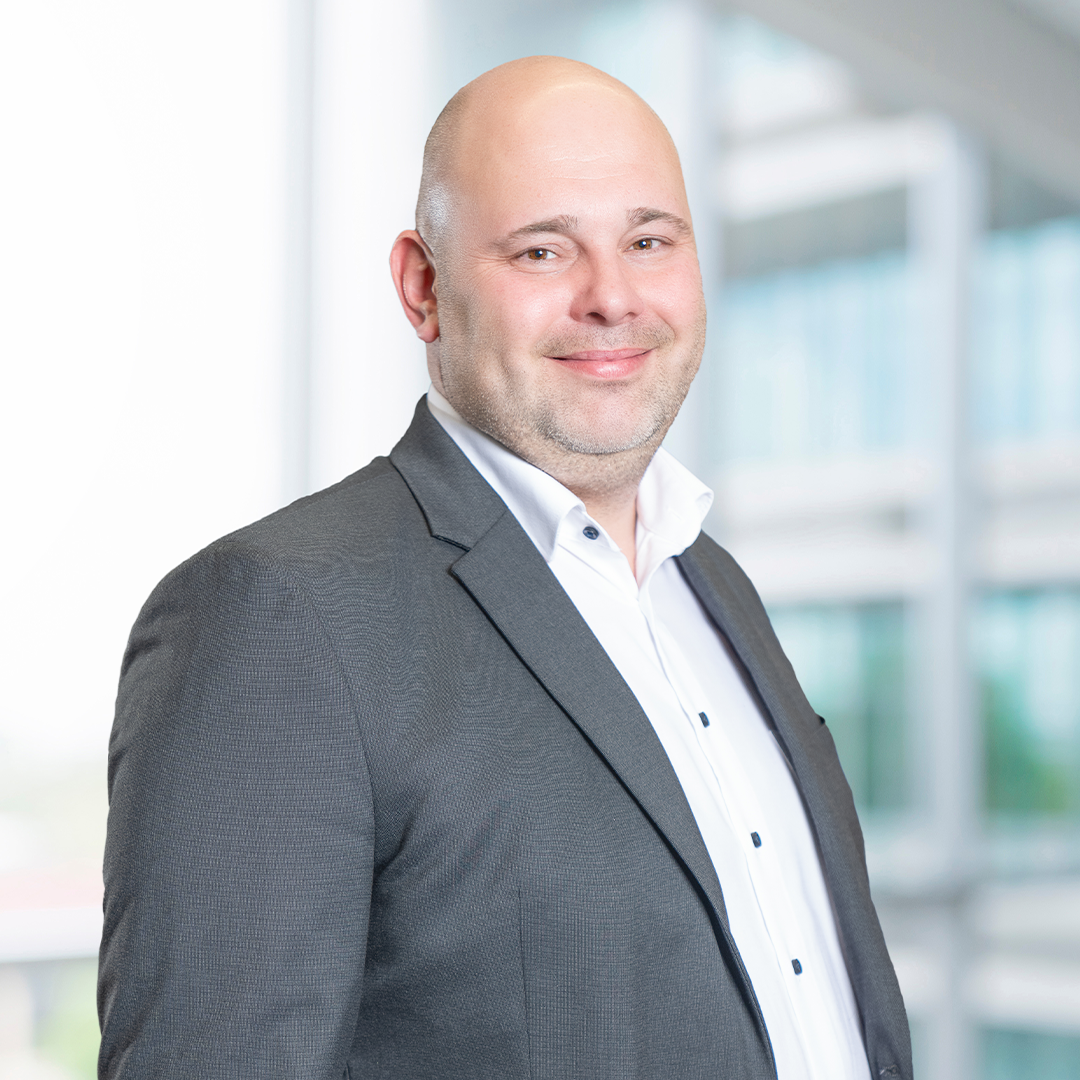 A smiling man with a bald head is wearing a dark blazer over a white shirt. He stands in a brightly lit environment, with large windows and a modern building visible in the background.