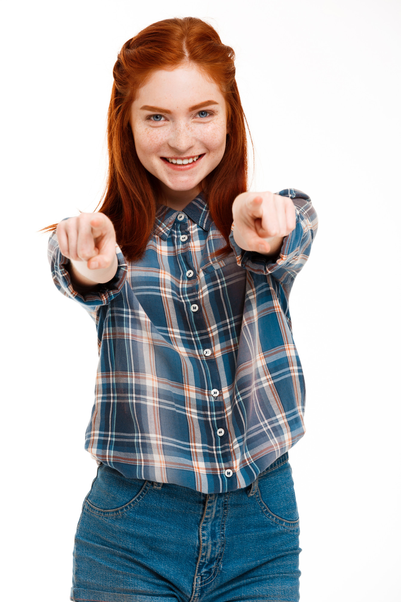 A girl with red hair and freckles smiles and points directly at the camera while wearing a plaid shirt and denim jeans.