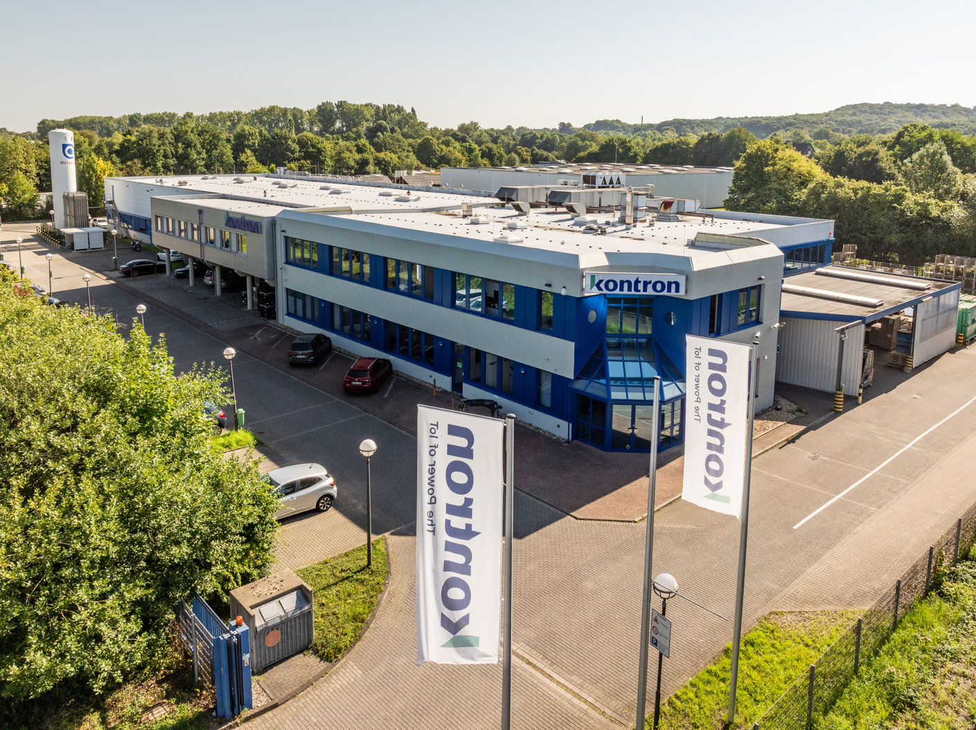 Ein modernes Firmengebäude mit dem Namen "Kontron" an der Fassade. Umgeben von grüner Landschaft und Parkplätzen. Sonniger Tag mit klarem Himmel. Banner mit dem Unternehmenslogo im Vordergrund.