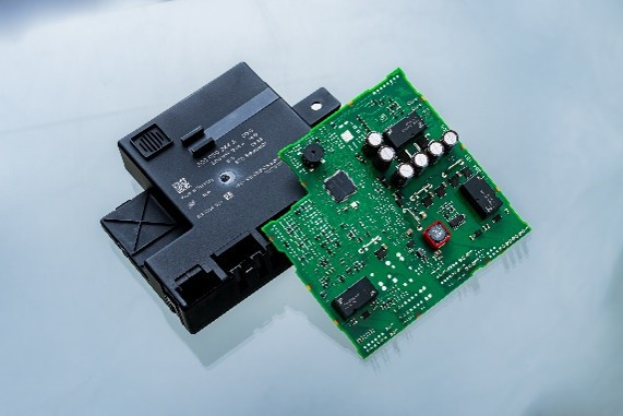 Eine grüne Leiterplatte mit verschiedenen elektronischen Bauteilen und eine schwarze, rechteckige Steuereinheit liegen auf einer glatten Oberfläche.