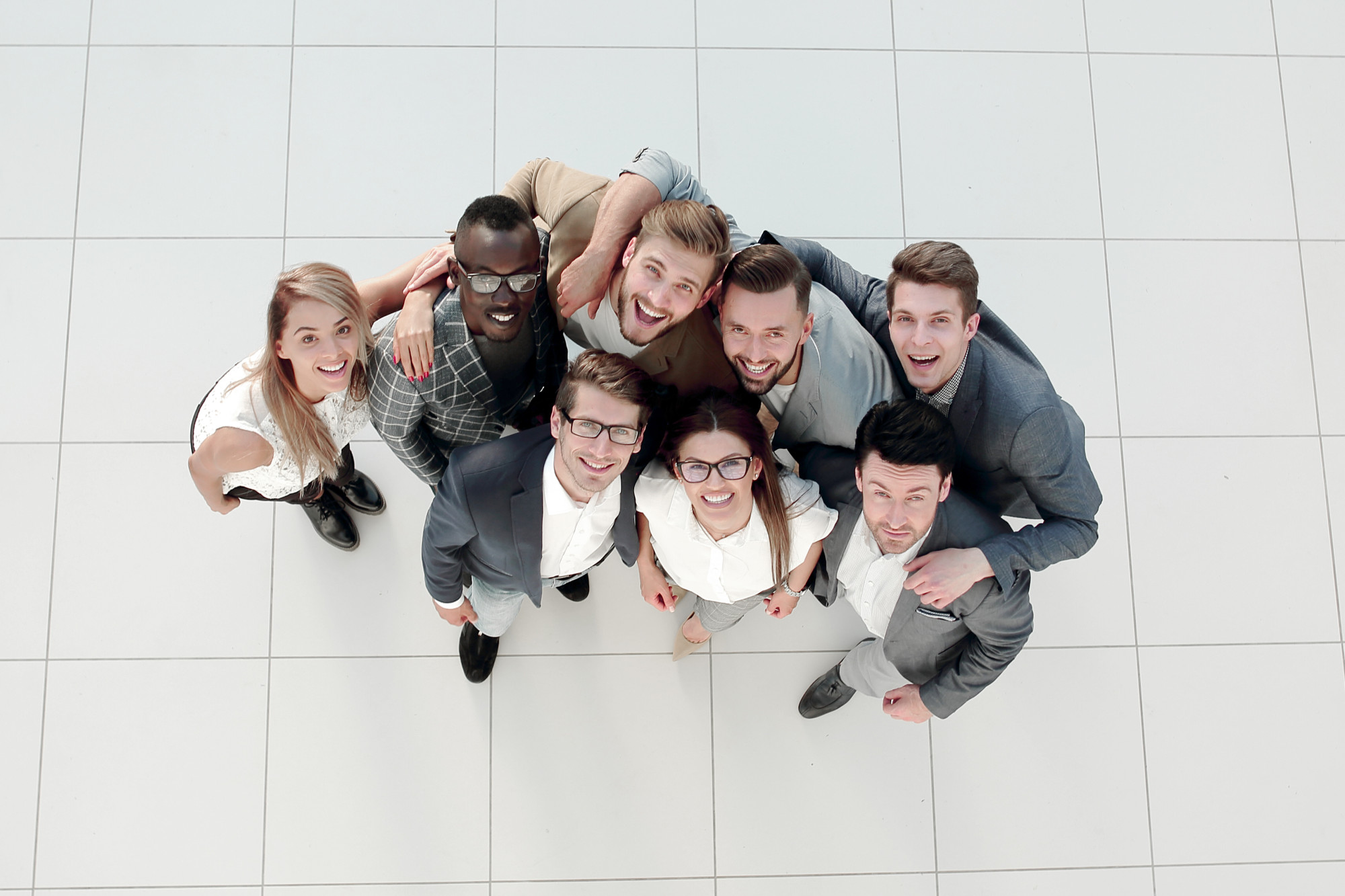 Eine Gruppe von neun Menschen steht zusammen und lächelt in die Kamera. Sie tragen formelle Kleidung und stehen auf einem hellen, gefliesten Boden.