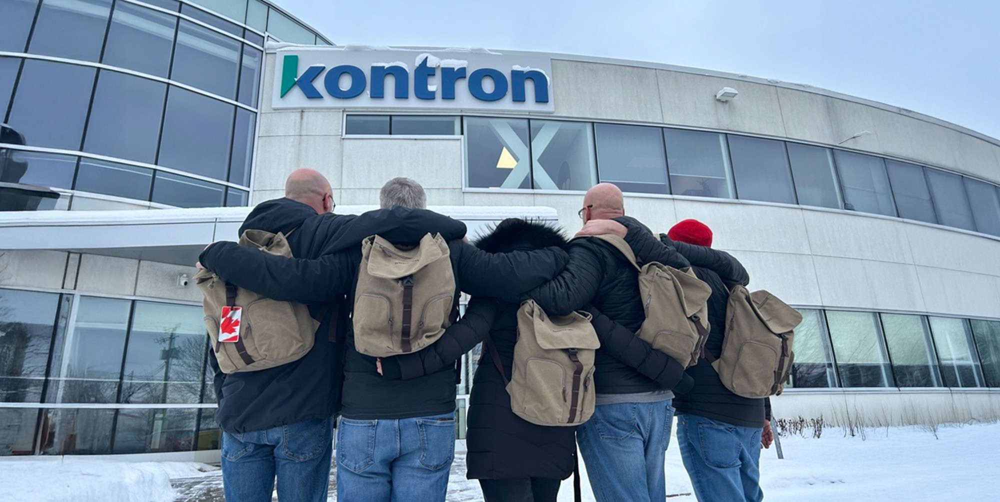 A group of six people stand with their backs to the camera, arms around each other, in front of a building displaying the "kontron" logo. They are wearing winter coats and backpacks, with snow on the ground and a cloudy sky above.