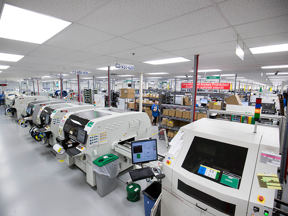 A large electronic manufacturing facility featuring multiple assembly machines and workstations. Workers are visible among neatly organized equipment and materials, with overhead lights illuminating the space. Signs indicate work areas and access restrictions.