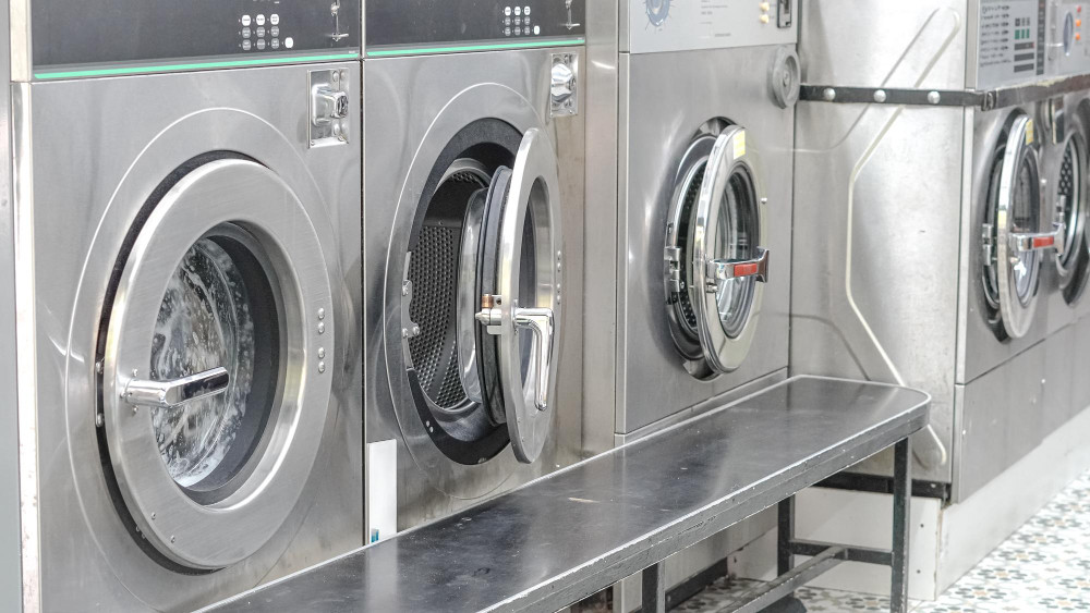 Several industrial washing machines lined up against a wall, featuring large circular doors and metal exteriors. A black countertop runs in front of the machines, with tiled flooring visible in the background.